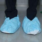 Front view of a person wearing blue disposable shoe covers over white shoes, standing on a tiled floor with black pants, and a green object partially visible on the right.
