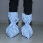 Person wearing high-rise disposable shoe covers tied at the ankles for medical or cleanroom use.