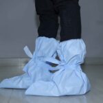 Side view of a person wearing light blue disposable shoe covers tied around the ankles, standing on a tiled floor with black pants.