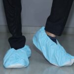 Side view of a person wearing light blue disposable shoe covers over white shoes, with one foot slightly lifted, standing on a tiled floor in dark pants.