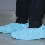 Side view of a person wearing blue disposable shoe covers over dark footwear, standing on a smooth tiled floor with black pants.