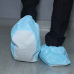 Rear view of a person wearing blue disposable shoe covers with white soles, standing on a tiled floor in black pants.