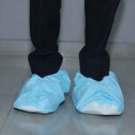Front view of a person standing on a tiled floor wearing light blue disposable shoe covers over black shoes and black pants.